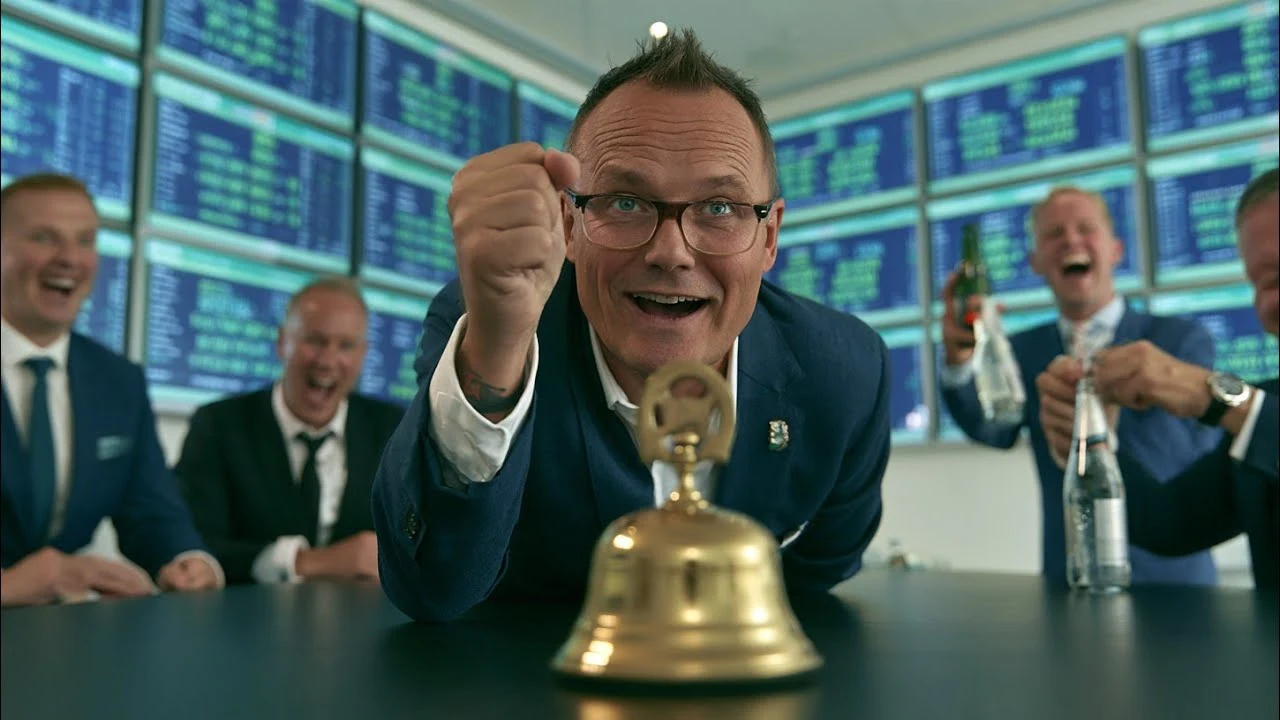 Suited man leaning toward a brass desk bell with cheering colleagues and market screens behind him